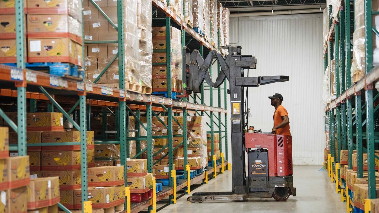 Warehouse worker in orange hard hat operating red pallet stacker between tall shelves of boxes