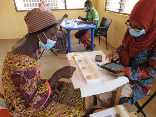 Two African women sit at a small table. The woman at left is looking at a white tray with yellow cubes in three wells. The woman at left takes notes on a tablet device.